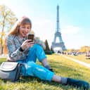 A joyful young blonde woman sitting on the grass in Paris, using her smartphone with the Eiffel Tower in the background.