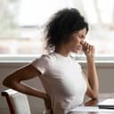 Close-up of a tired young woman at her desk, touching her lower back, experiencing pain.