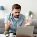 Close-up of an angered young man stressed by a computer malfunction, seated in his living room.