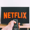A person holding a remote control aimed at a TV screen displaying the Netflix interface.