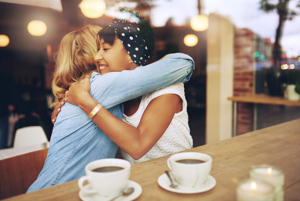 Two young women in a coffee shop hugging each other and smiling after making up 
