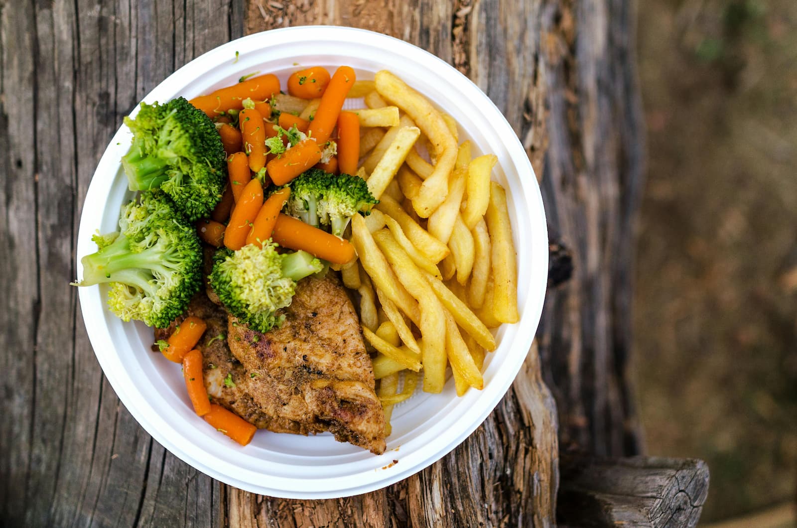 A white plate with chicken, fries, carrots and broccoli on a wooden table